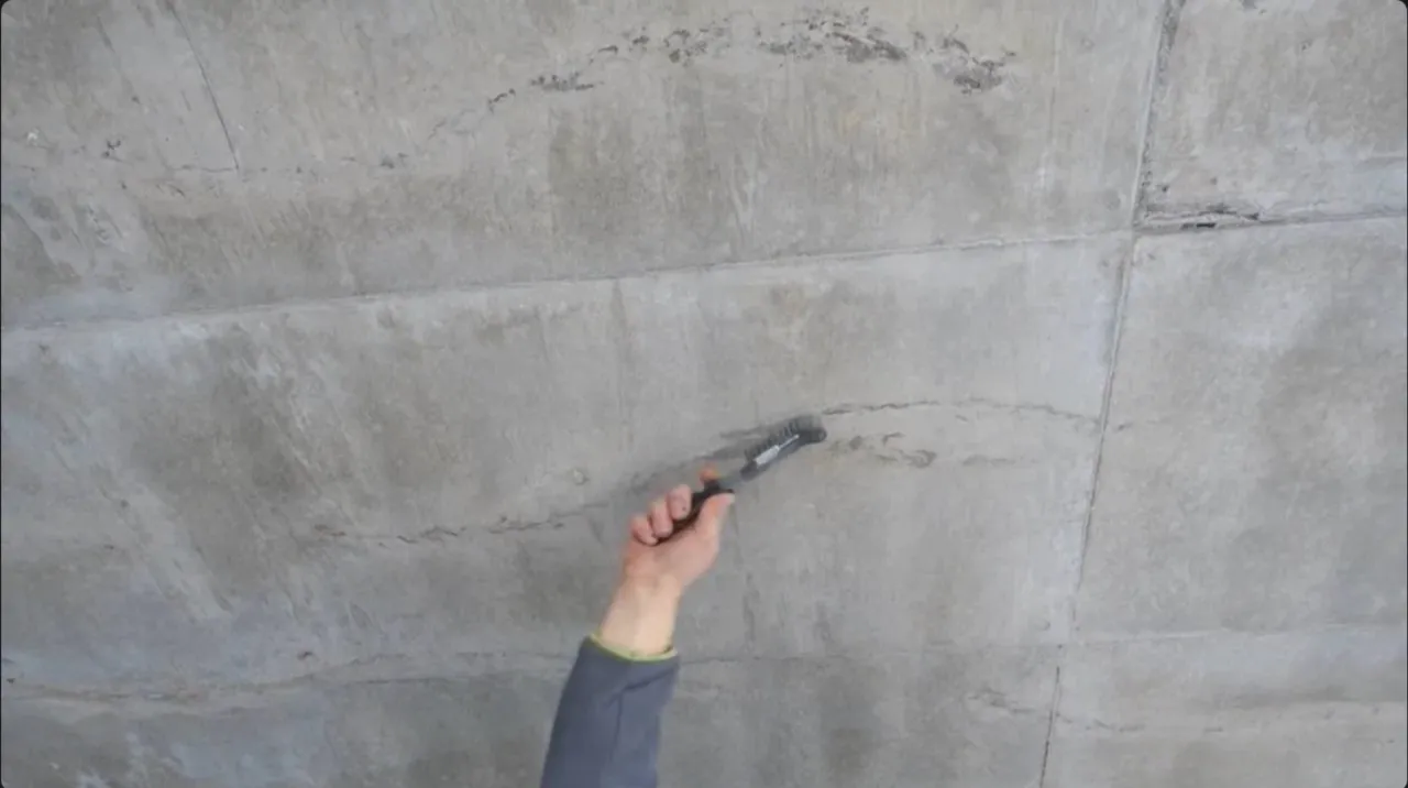 Hand mit Drahtbürste reinigt eine Sichtbetonwand mit Rissen und Arbeitsfugen