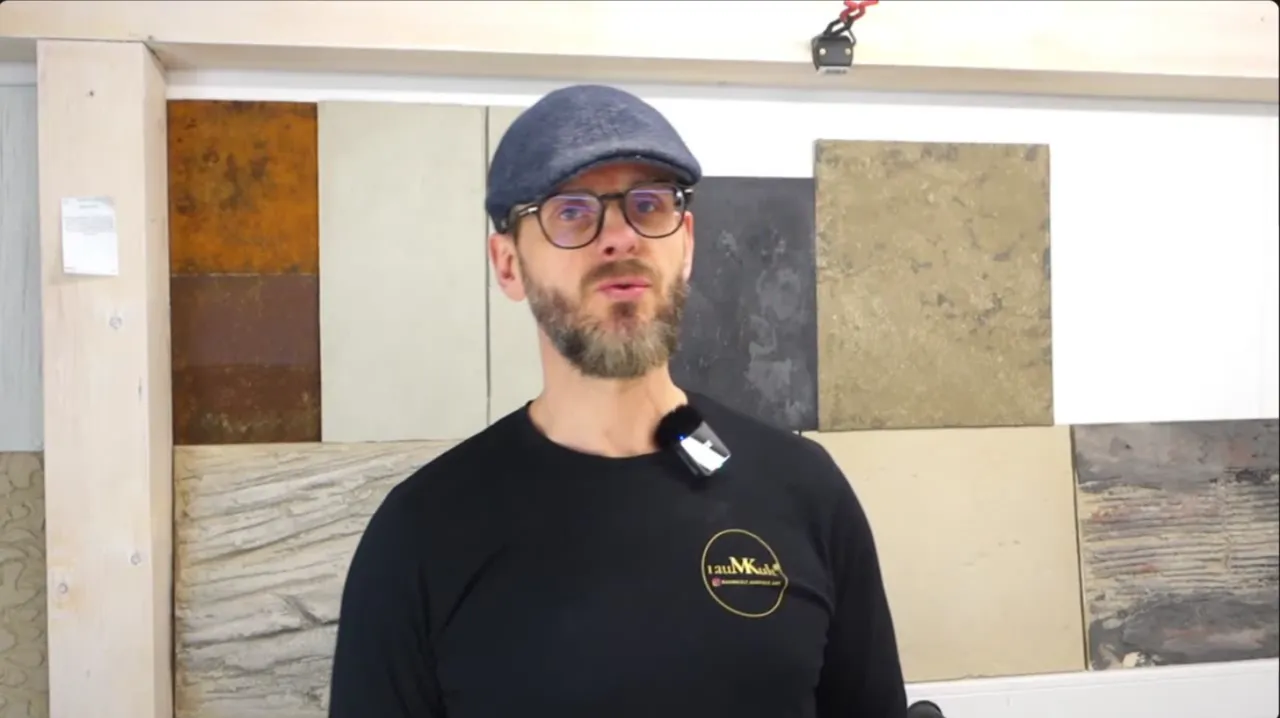 Kai Marienfeld mit Schiebermütze, Brille und schwarzem rauMKult-Shirt vor einer Wand mit Musterplatten in Beton- und Rostoptik