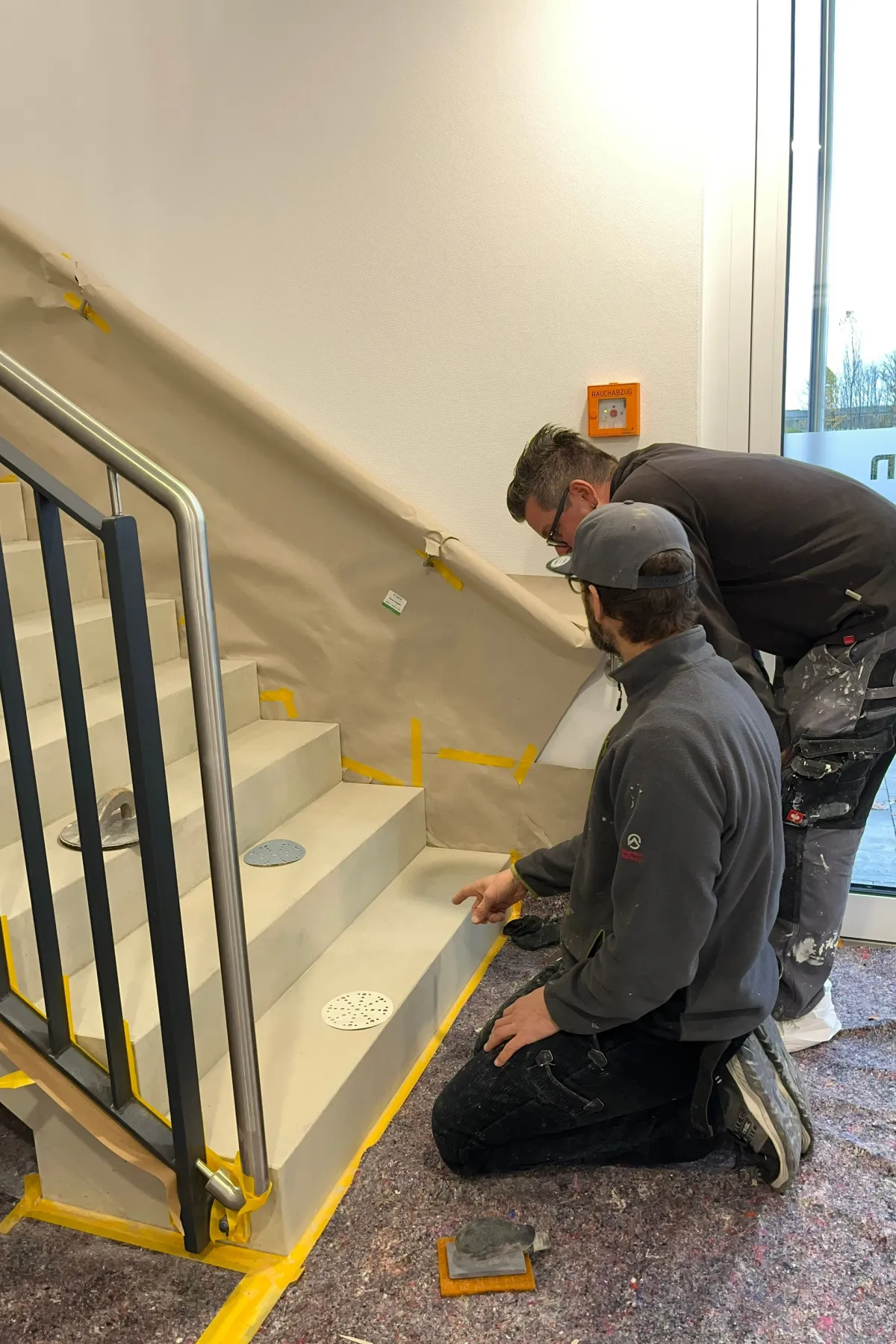 Zwei Handwerker prüfen bei einer Vor-Ort-Schulung eine frisch bearbeitete Betontreppe im Treppenhaus