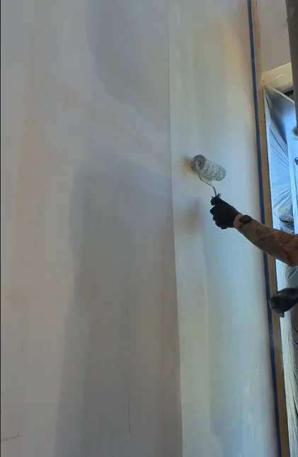 Eine behandschuhte Hand rollt mit einer Farbrolle das Gewebe auf einer hohen hellen Wand an, links daneben bereits die helle Fläche, rechts blaues Abklebeband und Schutzfolie