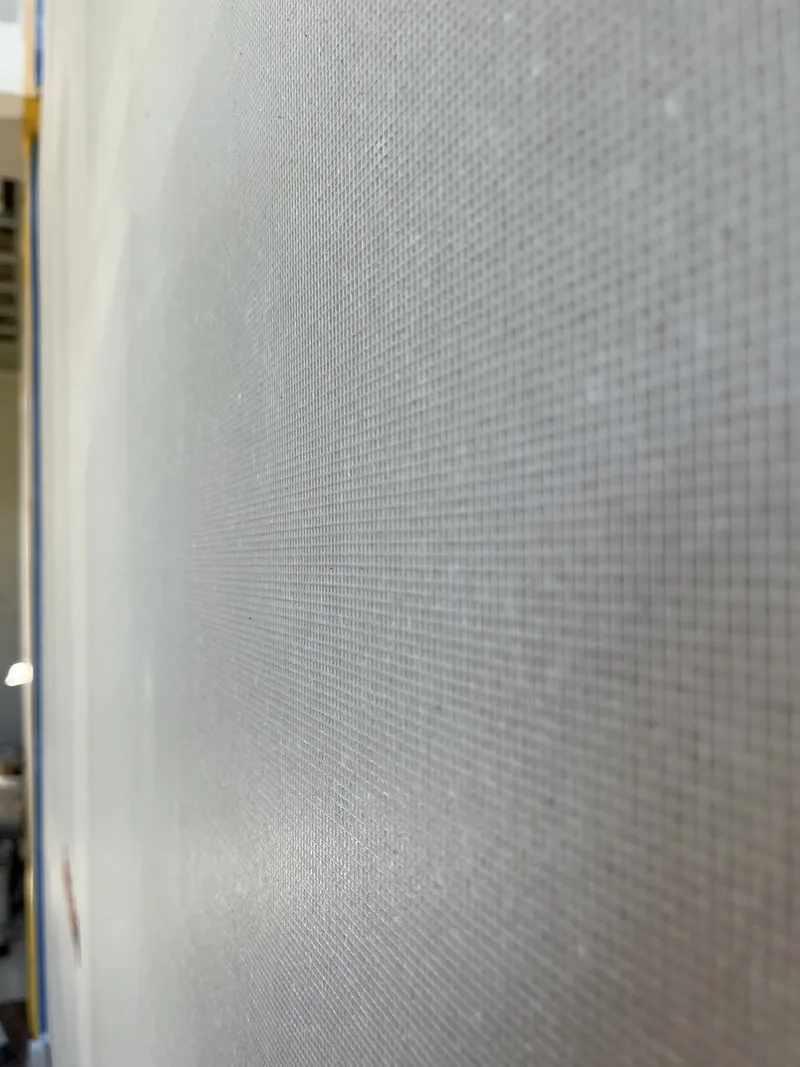 Feinmaschiges helles Gewebe liegt als gleichmäßiges Gitter auf einer Trockenbauwand, schräg von der Seite fotografiert, am linken Rand blaues Abklebeband