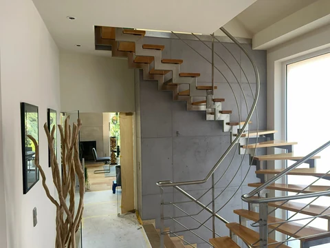 Treppenhaus mit Wand in Schalbetonoptik hinter einer freitragenden Treppe mit Holzstufen und geschwungenem Edelstahlgeländer