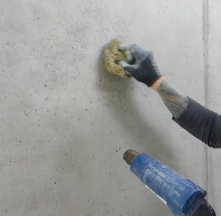 Hand mit Schwamm gleicht eine Sichtbetonwand farblich an, unten im Bild ein Heißluftgerät