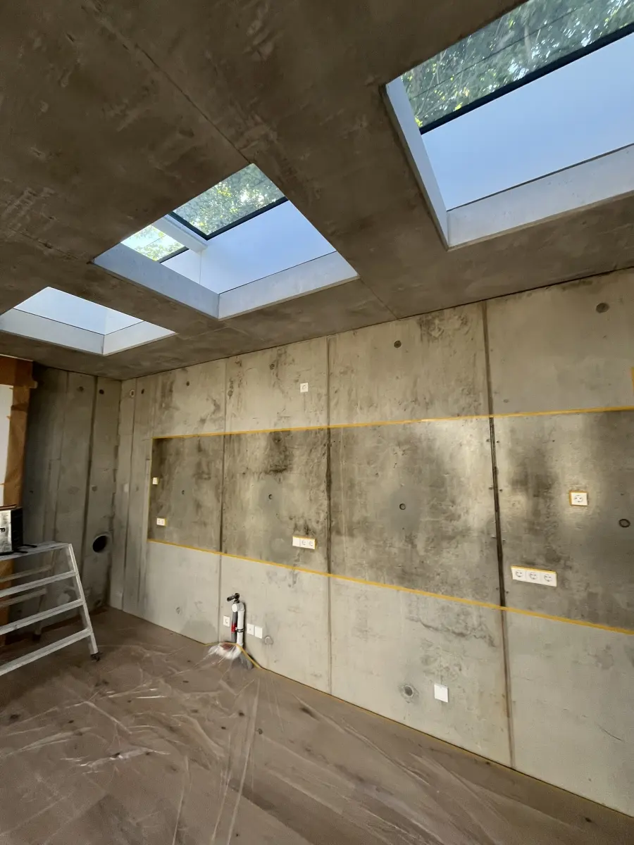 Sichtbetonwand unter einer Betondecke mit drei Dachfenstern: Ein mit gelbem Klebeband abgegrenztes Wandfeld ist nach der Mattversiegelung deutlich dunkler und kontrastreicher als die Wand daneben
