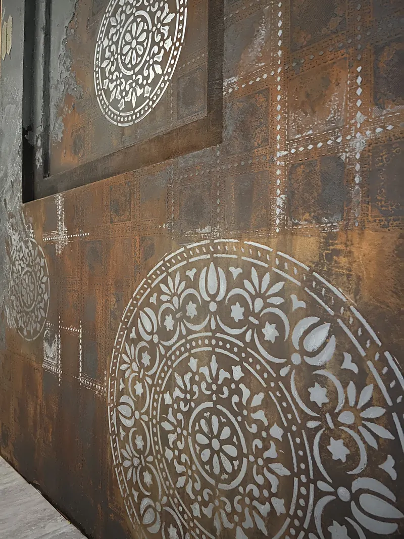 Wand in Rostoptik, schräg von der Seite fotografiert, mit weißen Ornamenten aus der Schablone: unten ein großes rundes Ornament, darüber ein Raster aus gepunkteten Linien, oben links ein eingelassenes Feld mit Rahmen und weiterem Ornament