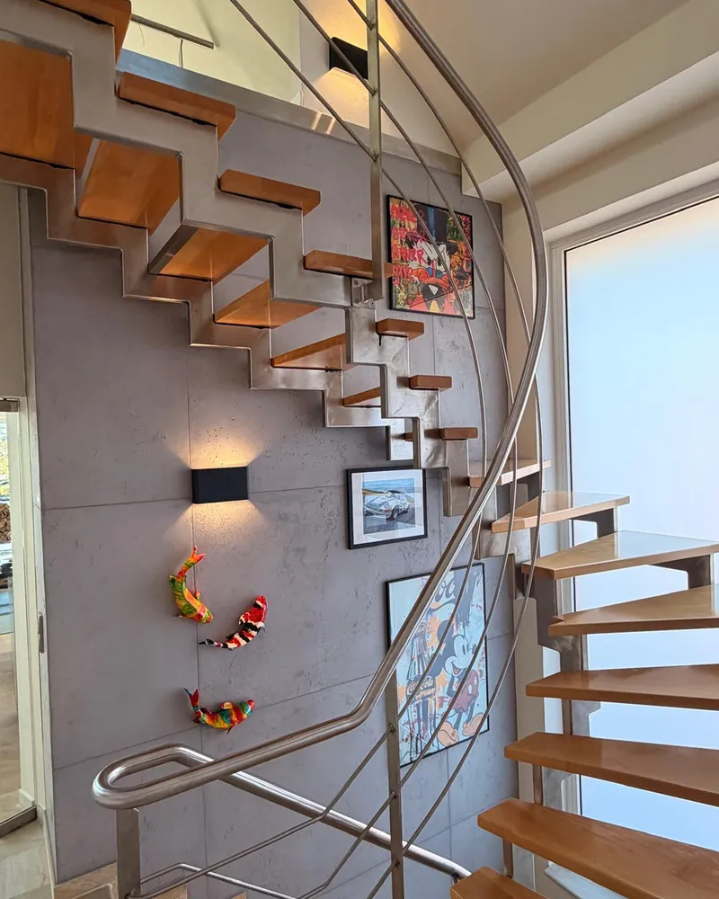 Treppenwand in Betonoptik mit Plattenraster hinter freitragenden Holzstufen, daran eine Wandleuchte, Bilder und bunte Figuren