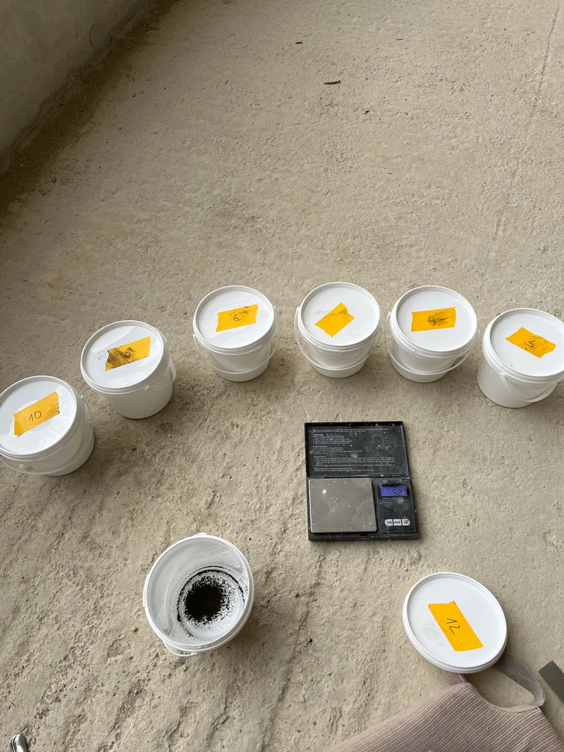 Mischbecher im Halbkreis um eine digitale Feinwaage auf dem Boden, ein Becher mit dunkler Mischung