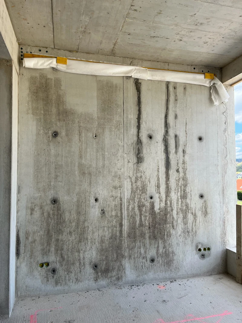 Große Sichtbetonwand im Rohbau mit dunklen Kiesnestern und aufgerissener Betonhaut über weite Teile der Fläche