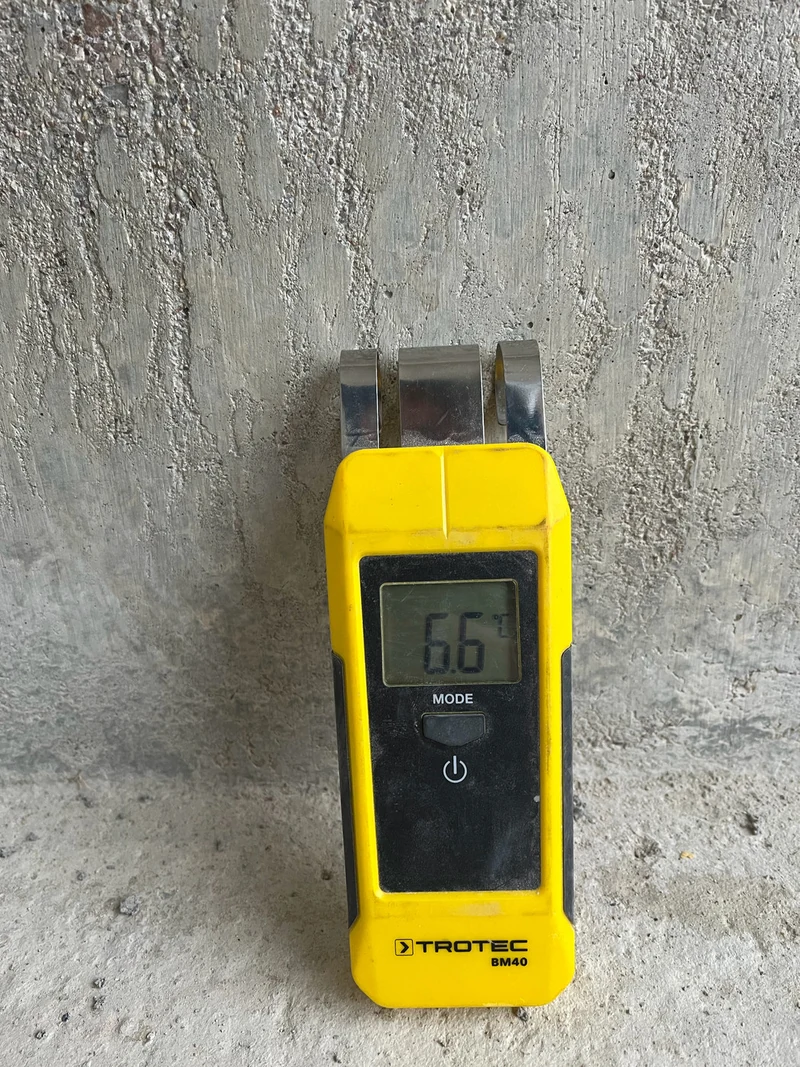 Gelber Feuchtemesser an einer Sichtbetonwand mit Brettstruktur, Anzeige im Display