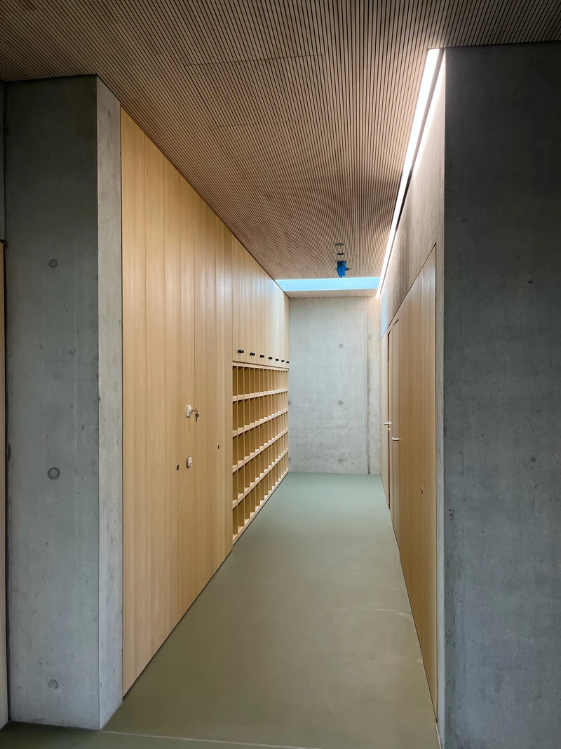 Schmaler Garderobenflur mit Schränken und Fächern aus Holz, vorne Stützen aus Sichtbeton direkt an den Einbauten, oben ein Lichtband