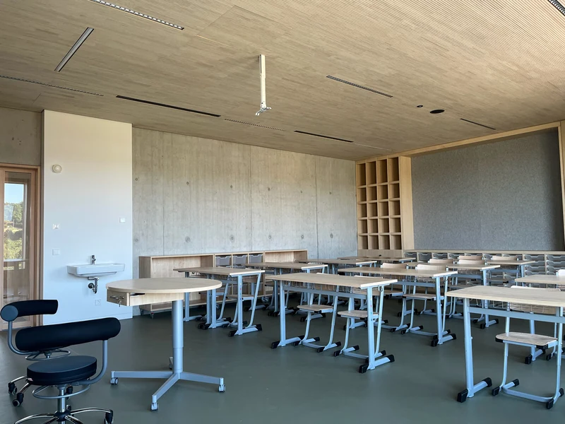 Klassenraum mit Tischen und Stühlen vor einer langen Sichtbetonwand, oben Holzlamellendecke, rechts ein Holzregal