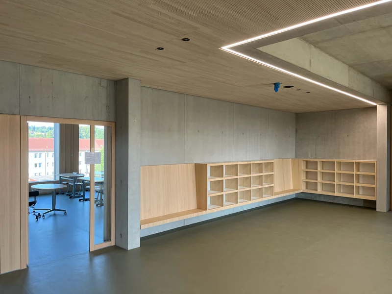 Raum mit Sitzbank und Fächern aus hellem Holz vor Sichtbetonwänden, darüber Holzdecke mit Lichtband, links der Blick nach draußen