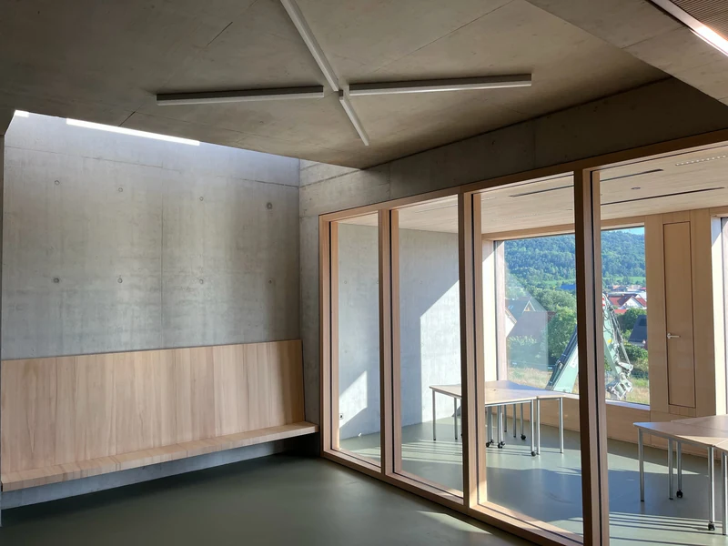 Blick aus dem Flur durch die Fenster der Trennwand in den Nachbarraum, oben Sichtbetondecke mit Kreuzleuchte, links Holzbank vor Sichtbeton