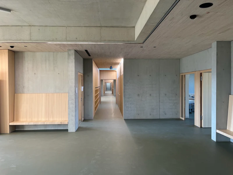 Flur mit Holzdecke und Betonunterzug, Sichtbetonwände links und rechts, dazwischen eine Holzbank in einer Nische