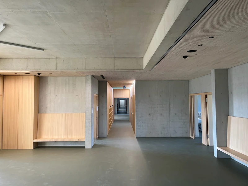 Langer Flur, rechts Holzdecke, mittig ein Unterzug aus Beton, links Betondecke und Sichtbetonwand, vorne Wände mit Holzeinbauten