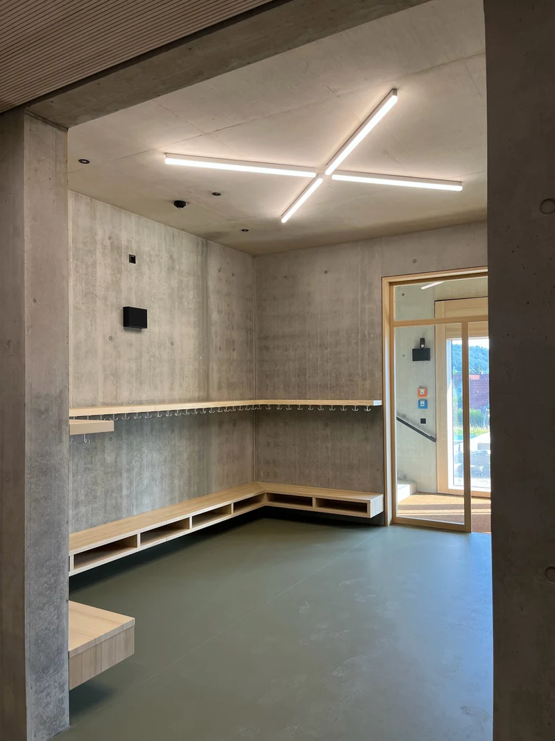 Eingangsgarderobe mit Bänken und Haken aus Holz, rundum Wände und Stütze aus Sichtbeton, darüber eine Leuchte in Kreuzform
