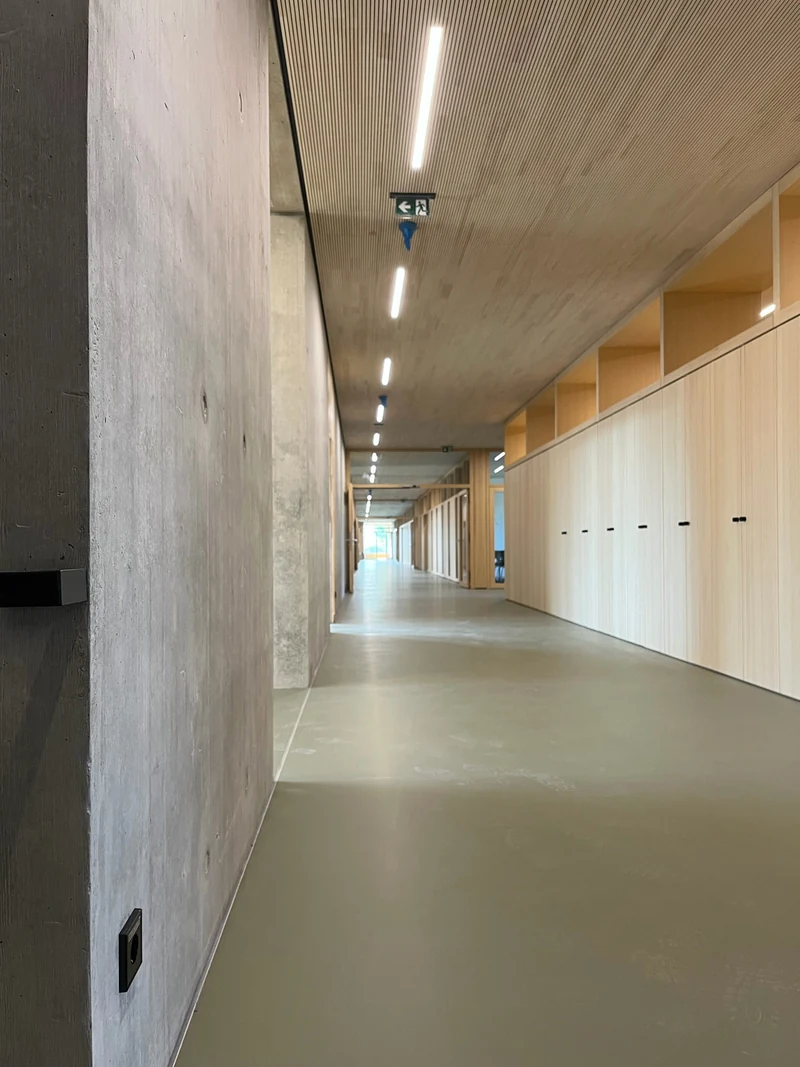 Langer Flur mit Sichtbetonwand links und Schrankwand aus Holz rechts, oben Holzlamellendecke mit durchgehendem Lichtband