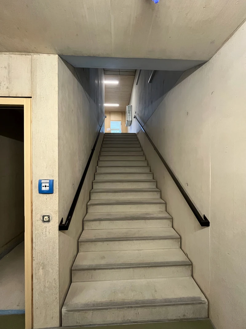 Gerade Treppe zwischen Sichtbetonwänden, schwarzer Handlauf, Stufen aus Beton, oben ein schmales Lichtband