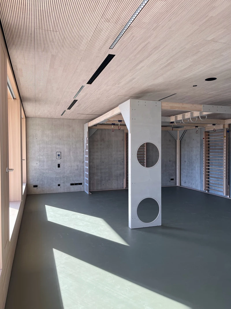 Sportraum mit Holzlamellendecke, hinten eine Sichtbetonwand, rechts und in der Mitte Einbauten aus Holz für die Kletterwand