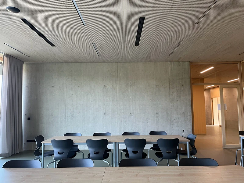 Klassenraum mit langen Tischen und schwarzen Stühlen vor einer großen Sichtbetonwand, darüber Holzdecke mit Leuchtstreifen