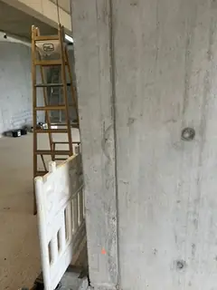 Betonstütze mit Brettstruktur im Rohbau, davor eine Leiter und eine weiße Absperrung, orange Markierung am Fuß