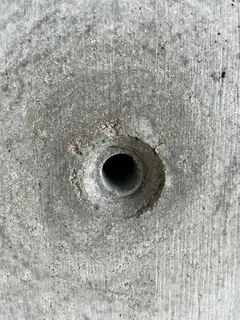 Spindelloch im Sichtbeton mit rauem, ausgebrochenem Rand und dunklem Hof ringsum, feine Brettmaserung