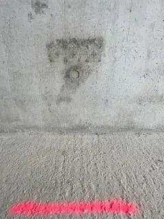 Sichtbetonwand mit dunklem, T-förmigem Fleck über einem Ankerkonus, am Wandfuß eine rote Markierung