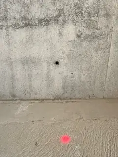 Sichtbetonwand mit einem Ankerloch über einem rauen Sockelbereich, am Boden ein rosa Markierungspunkt