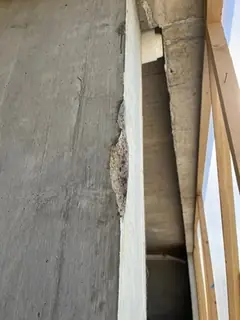 Betonkante einer Stütze mit kleiner Aufwölbung und Abplatzung, rechts Holzlatten und ein Rohr