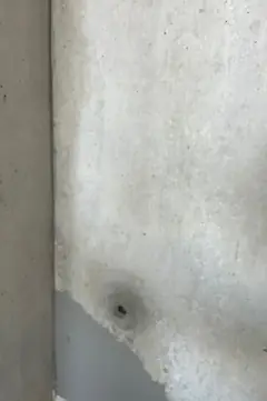 Abgeplatzte Betonhaut an einer Wand mit Ankerloch, darunter eine glatte, graue Fläche mit Kante