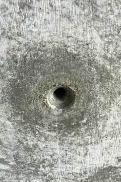 Kleines Spindelloch mit rauem Rand und dunklem Hof in heller, fleckiger Sichtbetonfläche