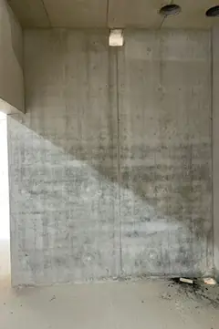 Sichtbetonwand mit diagonalem Lichtstreifen, oben eine weiße Leuchte, unten rechts Schutt am Boden