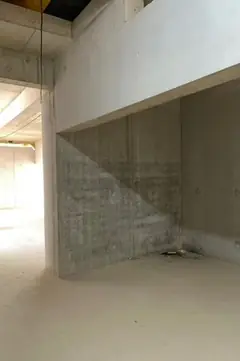 Blick in einen Rohbauraum mit Betonwand unter einem hellen Unterzug und einem offenen Durchgang links