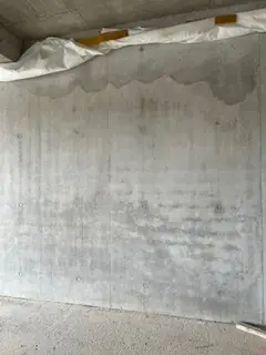 Sichtbetonwand mit wolkiger Oberfläche und weißer Abdeckung am oberen Rand, darunter ein Wasserrand