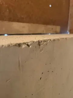 Obere Kante einer Betonfläche mit sandig-rauer, leicht ausgebrochener Zone, darüber braune Holzschalung