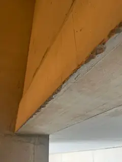 Betonkante eines Unterzugs mit krustig ausgebrochenem Rand rechts oben, daneben eine orange Wandfläche