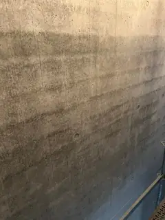 Betonwand mit waagerechten braunen Streifen in mehreren Tönen, unten rechts ein Gerüstteil