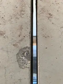 Abplatzung am Rand einer Betonplatte neben einer schmalen Fuge, auf der Fläche verstreute Sandkörner
