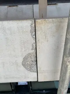 Zwei Platten mit Fuge, an der linken Plattenkante unten eine Abplatzung, rechts ein Gerüstrohr