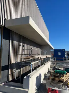 Fassade mit Holzlamellen, auskragendem Betonvordach und Gerüst, unten eine Betonbrüstung