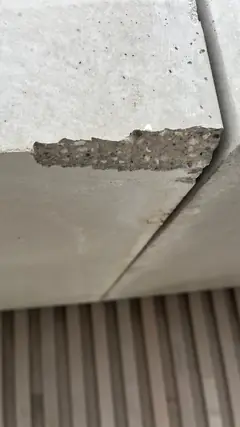 Abplatzung an einer Plattenecke mit grobem, freiliegendem Korn, im Hintergrund Holzlamellen