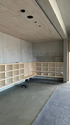 Raum mit Holzdecke, Betonwand und eingebauten Holzfächern, auf dem Boden ein Estrich mit Abdeckvlies