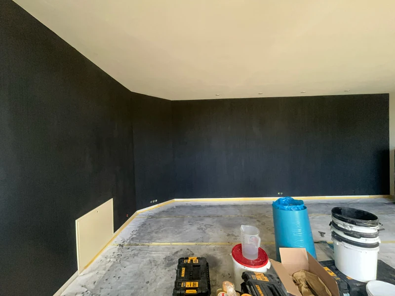 Schwarze Designwand mit Kalkstruktur in ganzer Länge im leeren Wohnraum, Abdeckung und Eimer am Boden