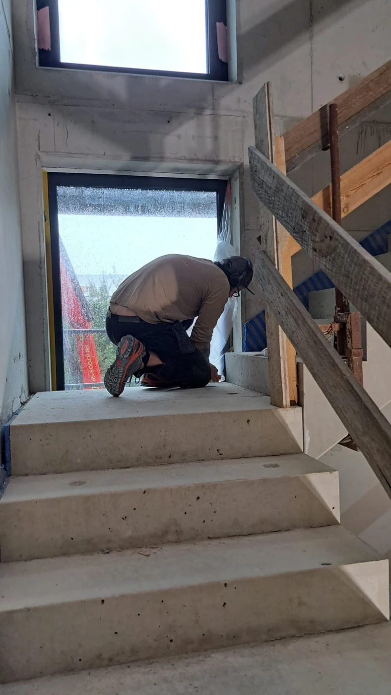 Handwerker kniet auf dem Podest eines Sichtbeton-Treppenhauses und klebt das Fenster vor der Betonkosmetik ab