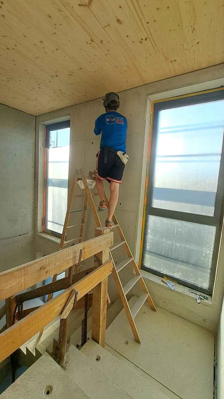 Handwerker auf der Leiter reinigt die Ortbetonwand an der Fensterseite im Treppenhauskopf unter der Holzdecke