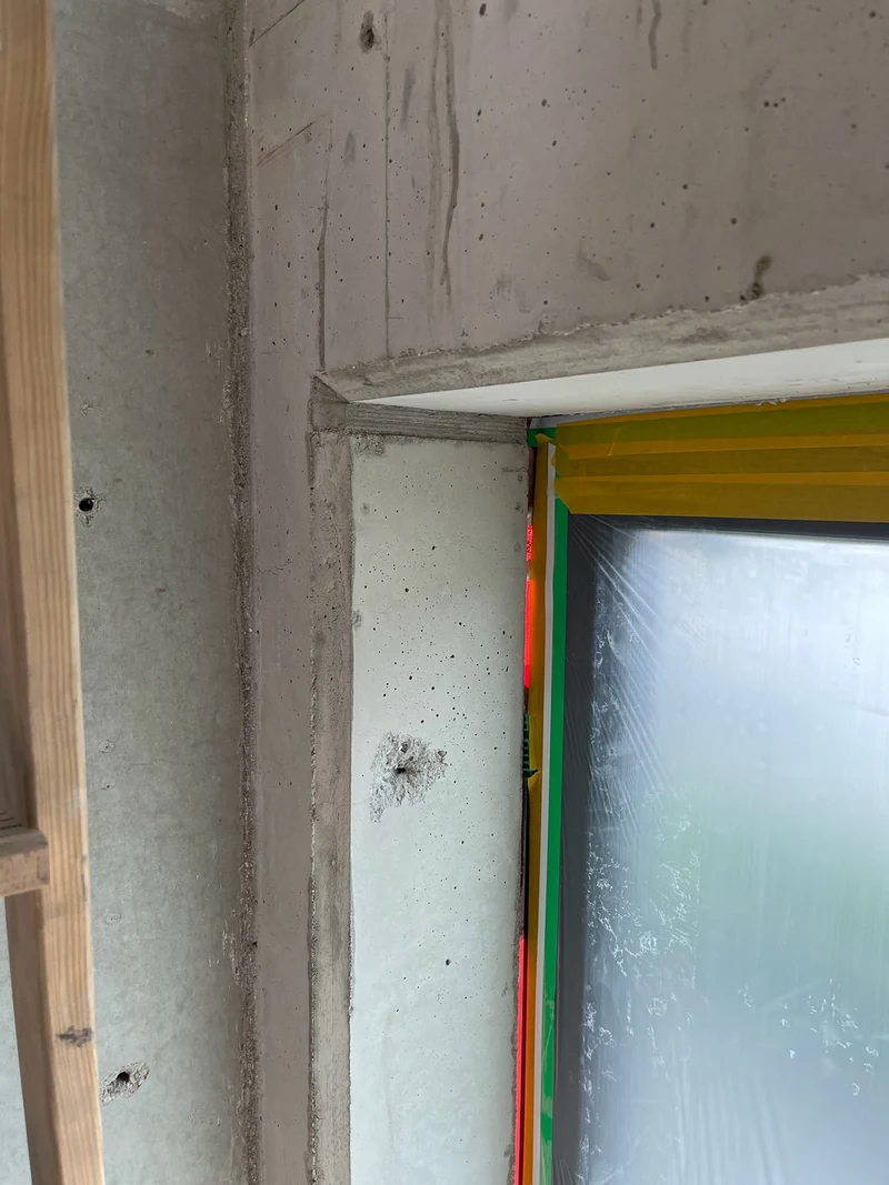 Vorher: Fensterlaibung mit Sturz und Bohrlöchern im Sichtbeton-Treppenhaus, das Fenster mit Schutzfolie abgedeckt
