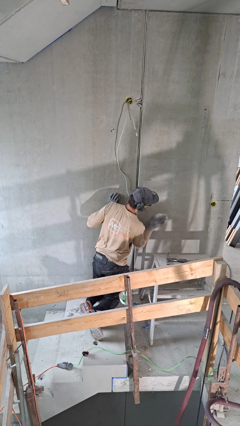 Weite Aufnahme im Sichtbeton-Treppenhaus: Handwerker kniet am Wandfuß und säubert die Betonwand mit Schleifpapier