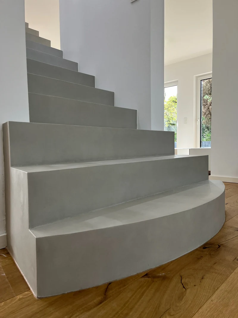 Runde Antrittsstufe aus Beton Ciré von der Seite, dahinter die Stufen und ein Fenster, Holzboden davor
